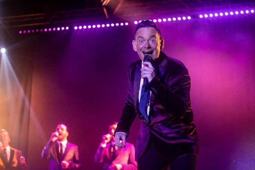 A person in a dark suit holding a microphone, standing on a stage with purple lighting. 2