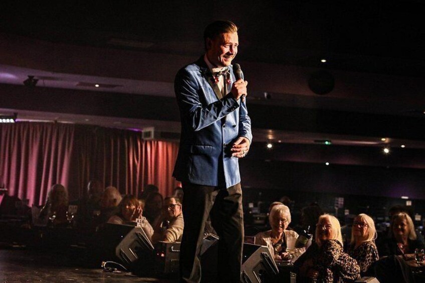 A person standing on a stage with a microphone, addressing an audience seated in rows. 2