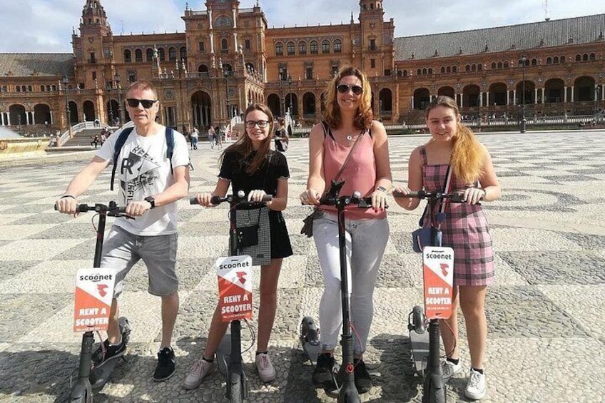 Quatre personnes sur des scooters, portant des panneaux indiquant « location de scooters », se trouvent devant un grand bâtiment historique. 2