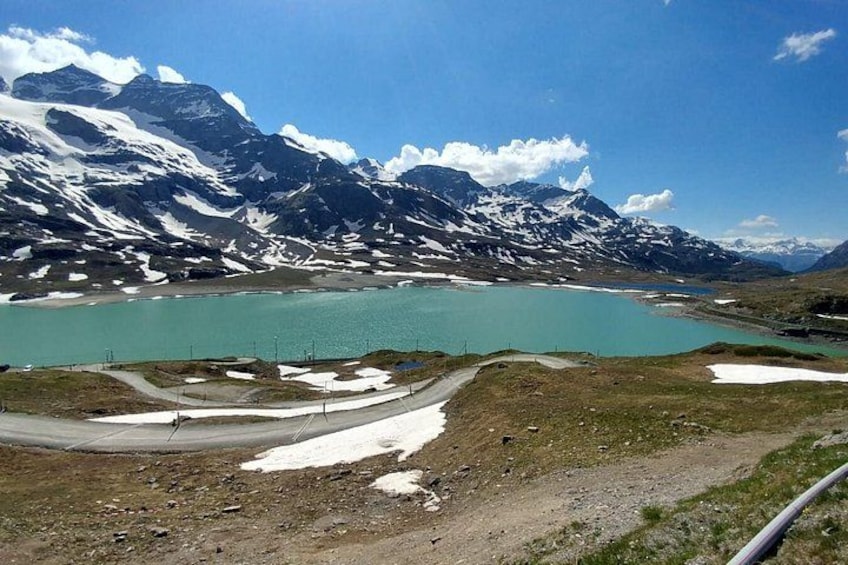 A mountainous landscape with a turquoise lake, snow-capped peaks, and a dirt road. 3