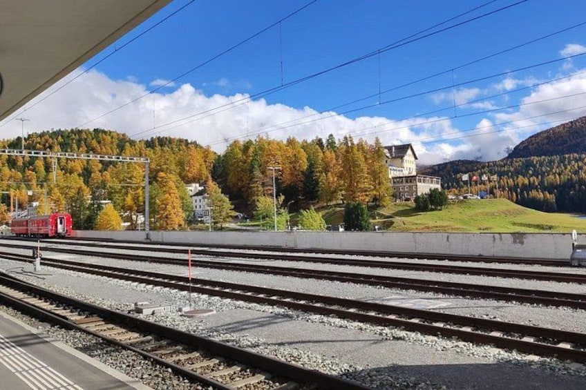 A railway station with multiple tracks, a red train, and a mountainous landscape with autumn-colored trees. 2