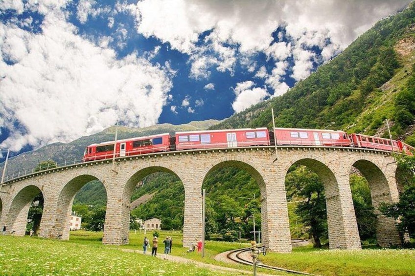 A red train on a stone bridge with arches, surrounded by greenery and mountains. 4