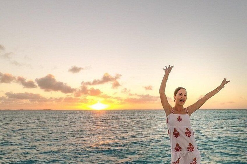 A person standing on a beach with arms raised, facing a sunset over the ocean. 1