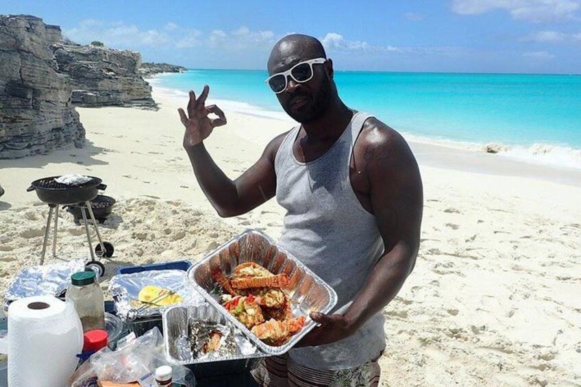 A person holding a tray of food on a beach with a grill and a rocky cliff in the background. 1