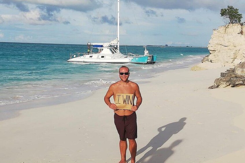 Una playa con una persona usando una camiseta con la frase 'I WIN', un bote y un acantilado rocoso. 2