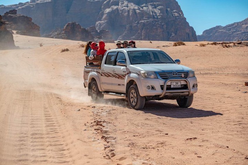 A white pickup truck driving on a sandy road in a desert with rocky mountains in the background. 4