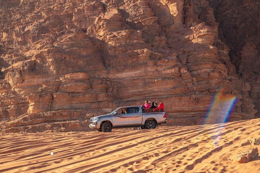 A white pickup truck with passengers on a sandy terrain with rock formations in the background. 5