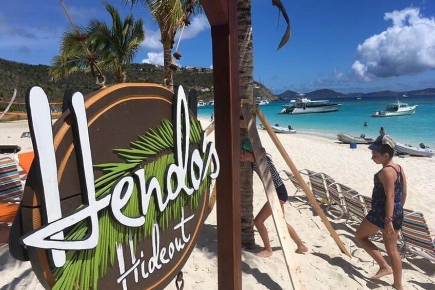A beach with a sign reading "Lendos Hideout," palm trees, and boats in the water. 1