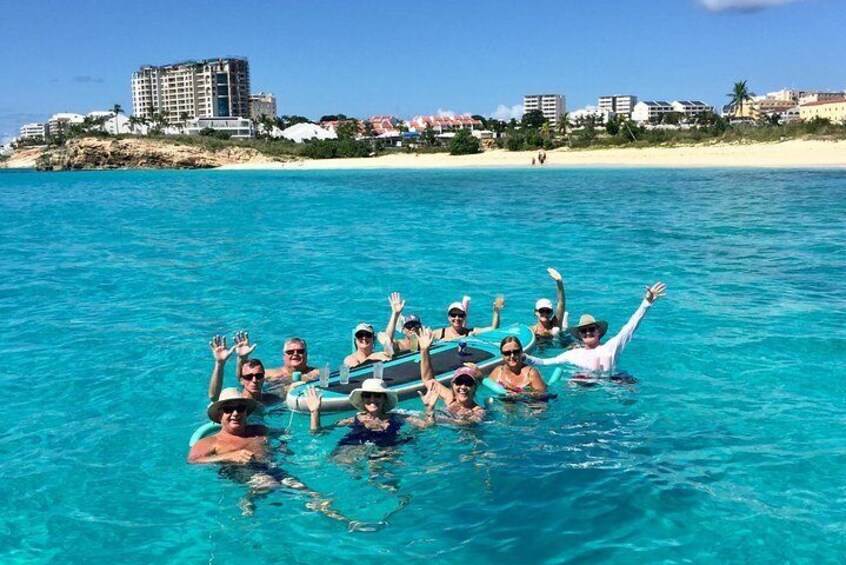 Eine Gruppe von Menschen treibt auf einem Boot in türkisfarbenem Wasser, im Hintergrund ein Strand und Gebäude. 3