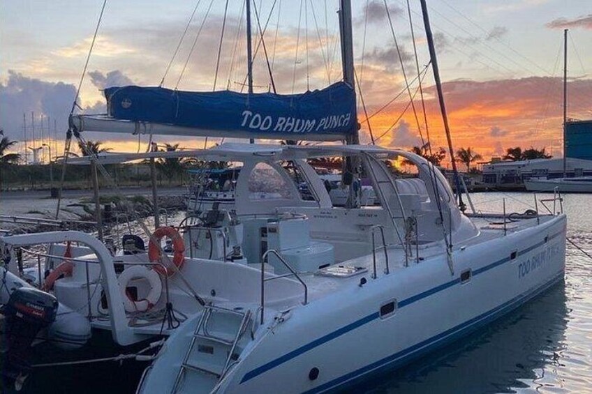 A white yacht docked at a marina with a blue canopy labeled 'TOO RHUM PUNCH' and a calm sunset in the background. 5