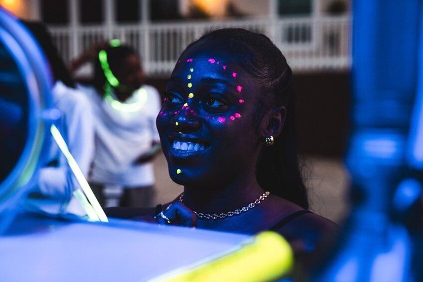 A person with glowing dots on their face, a chain necklace, and a blurred background with a railing. 2