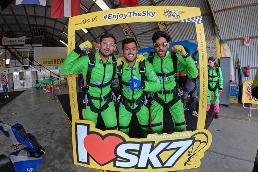 En gruppe mennesker i grønne dragter poserer foran et baggrund med teksten 'I ♥ SK7' og hashtagget '#EnjoyTheSky'. 3