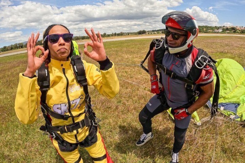 Two individuals in parachute gear standing in a grassy field with a clear sky. 3