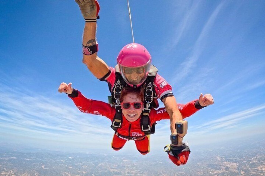 A person skydiving with a parachute, wearing a helmet and harness, against a clear sky and landscape below. 1