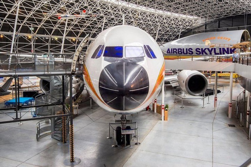 A large airplane with 'AIRBUS SKYLINK' written on its side, displayed indoors with a metal framework and other aircraft in the background. 4