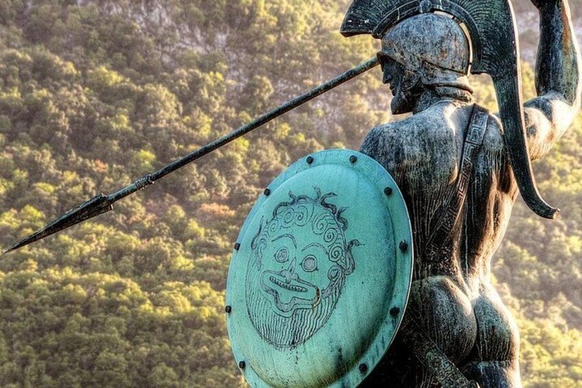 A statue of a warrior holding a spear and shield, set against a forested hillside. 2