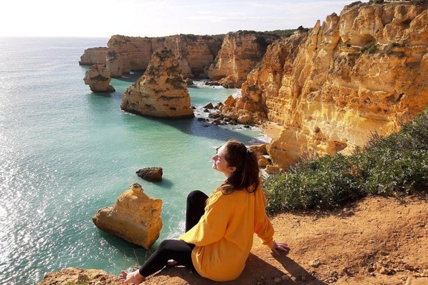 A person sitting on a cliff overlooking a coastal landscape with distinctive rock formations and clear blue water. 3