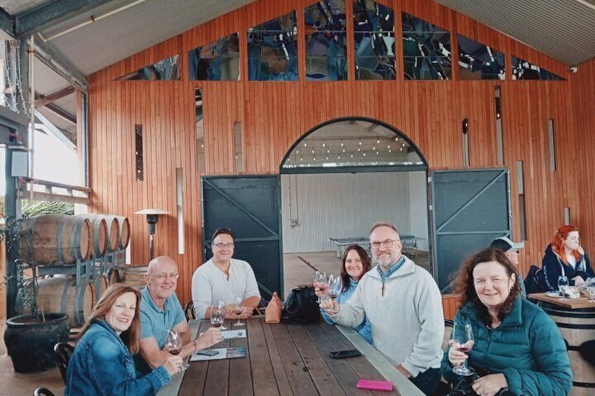 A group of people sitting at a wooden table outdoors, with wine glasses in hand, under a large wooden structure with a curved roof. 5