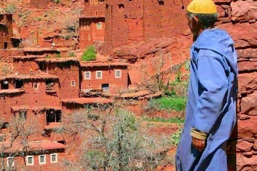 A person in traditional attire standing in front of a red rock landscape with terraced buildings. 3