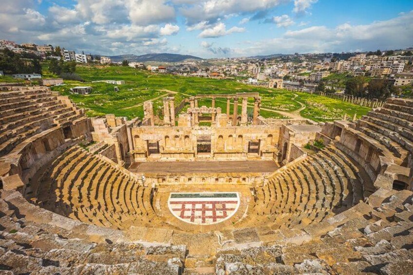 An ancient amphitheater with stone seating and a central stage, surrounded by green fields and modern buildings in the background. 4