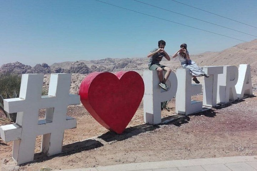 Dos personas sentadas sobre letras blancas grandes que deletrean 'PETRA', con un corazón rojo en el centro. 3