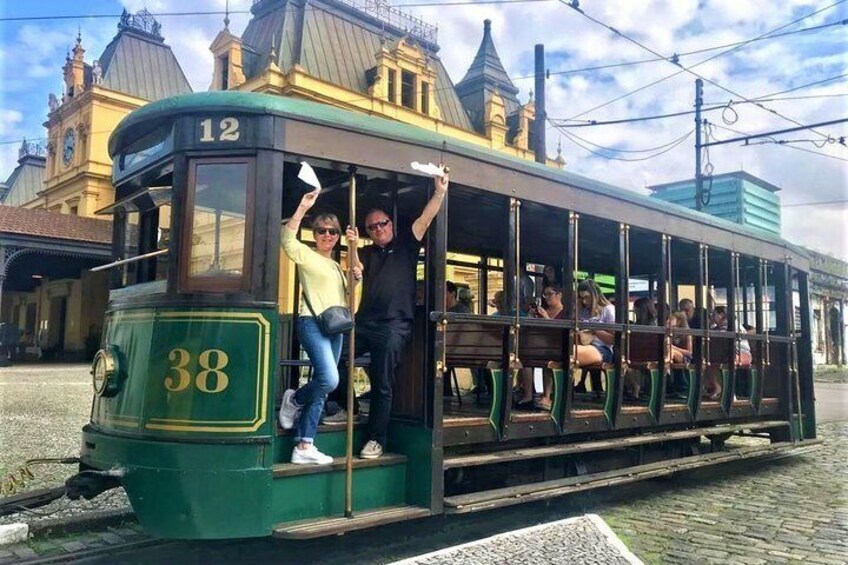 A green tram with the number 38 on the front, passengers inside, and a building with a clock tower in the background. 1