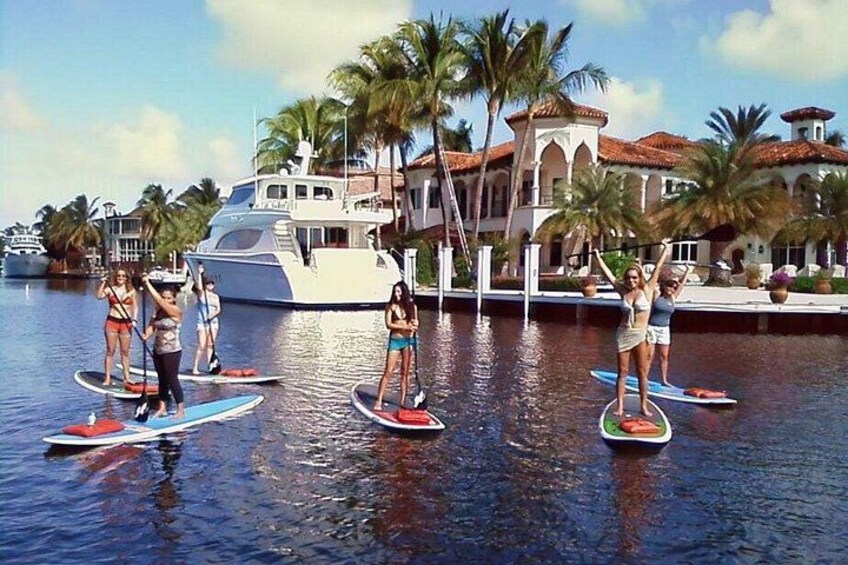 People paddleboarding on a calm body of water with a yacht and palm trees in the background. 5
