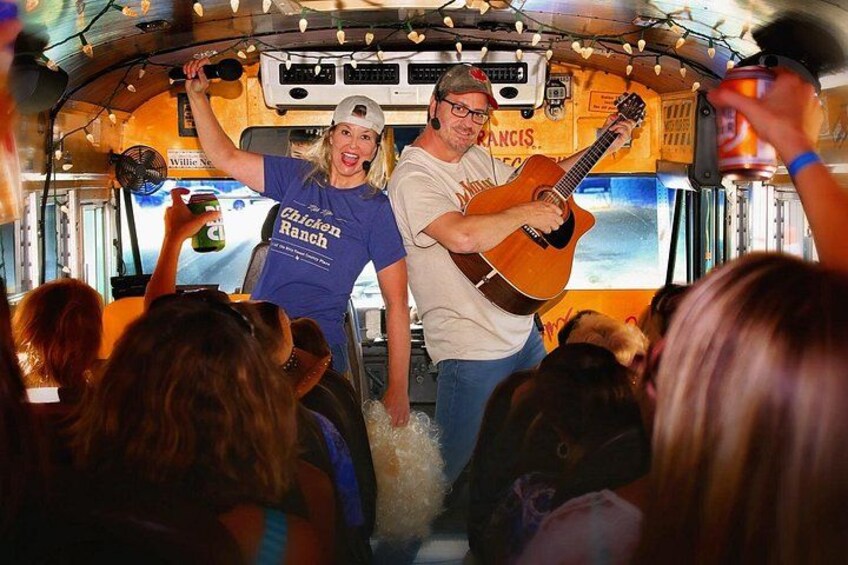 Een businterieur met twee mensen, waarvan één een gitaar bespeelt en de ander een microfoon vasthoudt, omringd door passagiers. 5
