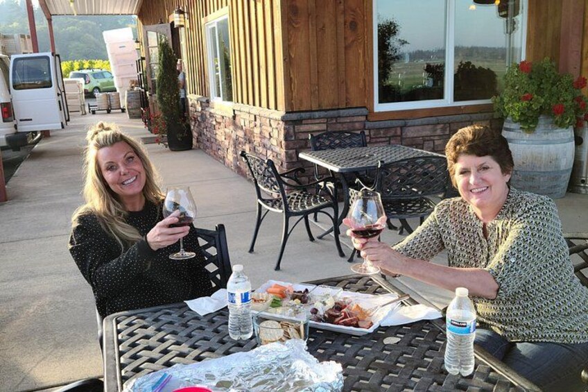 Two people sitting at an outdoor table with food and drinks, a building with a wooden facade in the background, and a parked van. 3