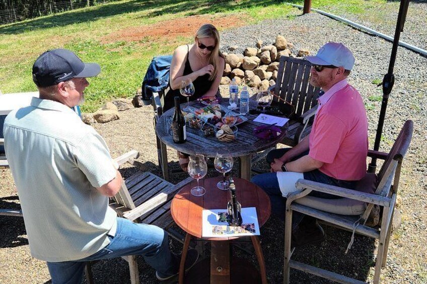 Tres personas sentadas en una mesa redonda de madera al aire libre, con una botella de vino, una botella de agua y varios alimentos. 5
