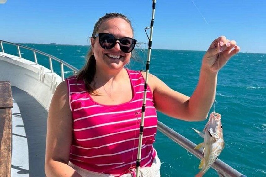 Eine Person hält einen Fisch in einem Boot, im Hintergrund das Meer und eine Reling. 2