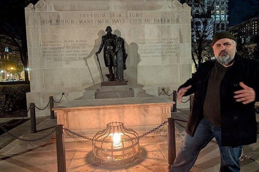 A statue with a plaque, a man standing beside it, and a chain barrier. 2