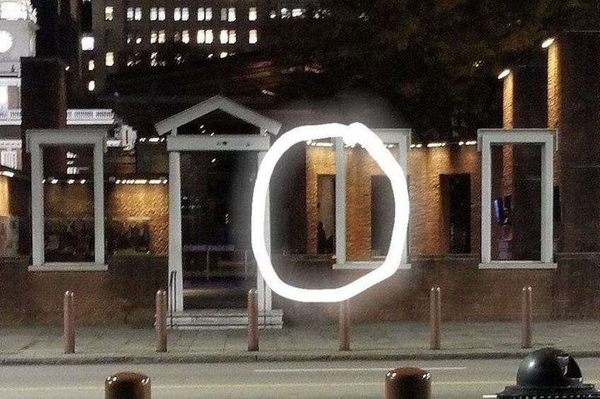 A nighttime urban scene with a white circular light installation in the foreground, a building with a triangular roof in the background, and a street with bollards. 5