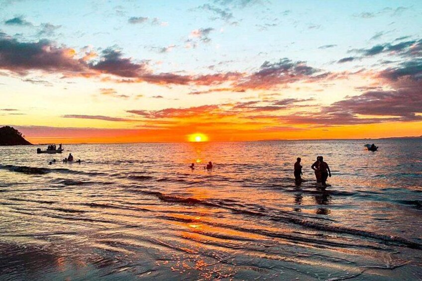 A sunset over a body of water with people wading and boats in the distance. 3