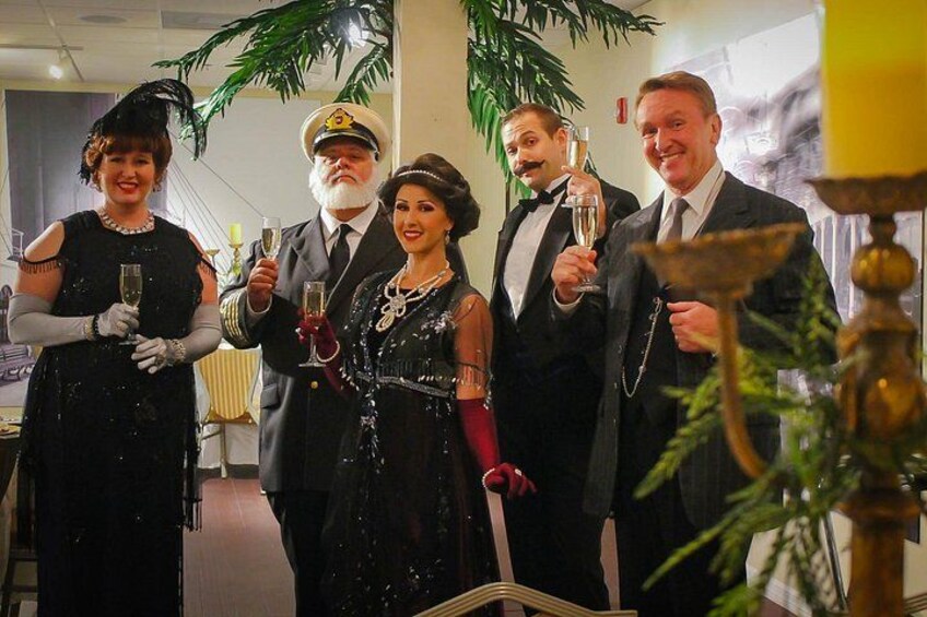 A group of people in formal attire holding drinks, standing indoors with a plant and a table in the foreground. 5