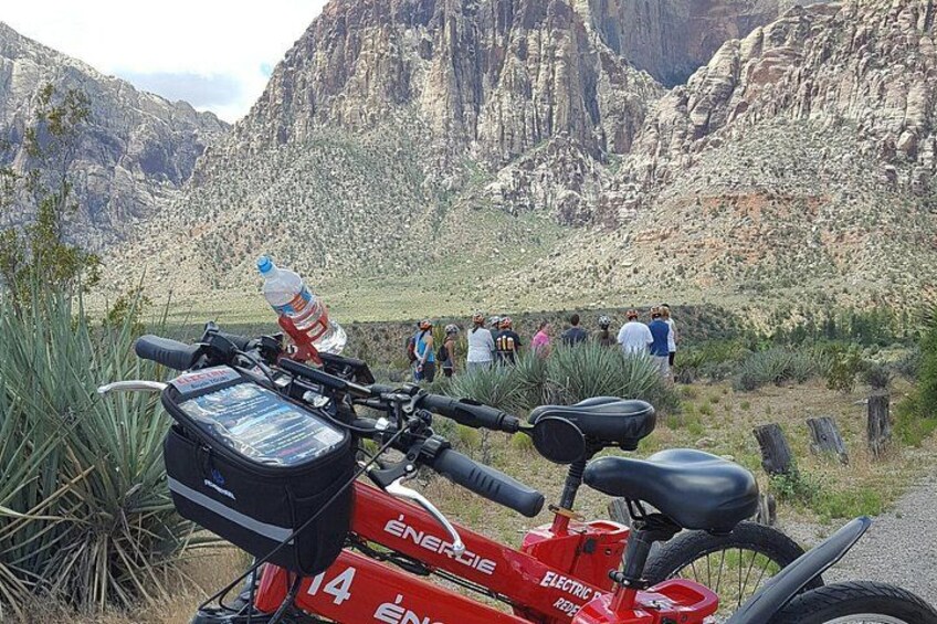 Une trottinette électrique rouge, équipée d’une bouteille d’eau et d’une carte, est garée dans une zone montagneuse, avec des personnes en arrière-plan. 2