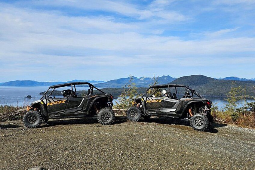 Two black off-road vehicles parked on a gravel road with a scenic view of mountains and a lake in the background. 1