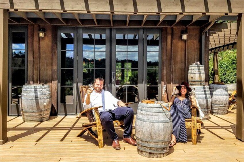 Une terrasse en bois avec des personnes assises sur des chaises, de grands tonneaux en bois et une structure en forme de pergola. 1