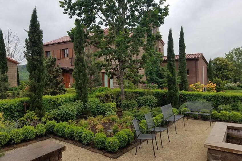 Une cour agrémentée de haies soigneusement taillées, un espace aménagé avec des chaises, et un bâtiment surmonté d’un toit recouvert de tuiles rouges. 2