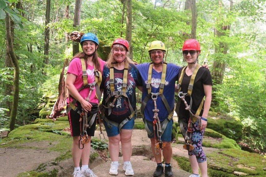 Four people in a forested area with hiking gear, wearing helmets and harnesses, standing on a trail. 4