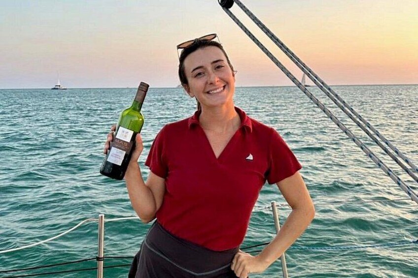 Une personne tenant une bouteille de vin sur un bateau, avec l’océan et un voilier au loin en arrière-plan. 5