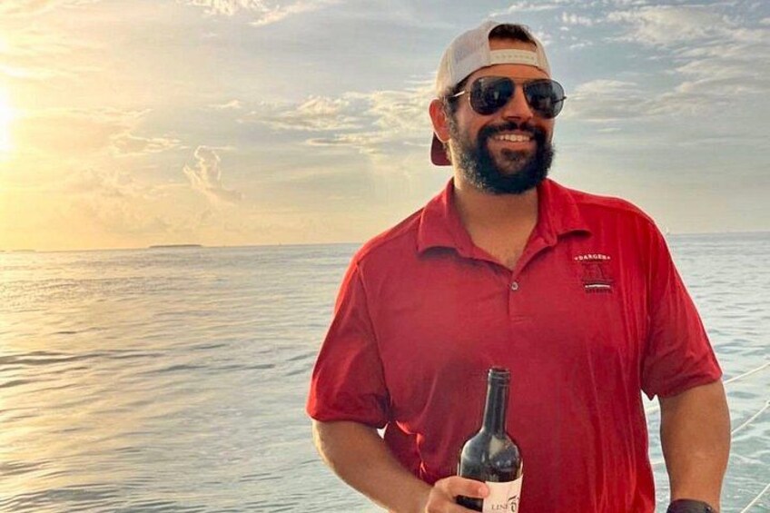 Une personne vêtue d’une chemise rouge, tenant une bouteille, se tient sur un bateau avec un coucher de soleil en arrière-plan. 2