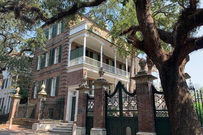 Un edificio histórico de ladrillo con un balcón, un árbol grande en primer plano y una entrada con reja. 4