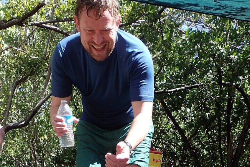 Een persoon vasthoudt een waterfles, draagt een blauw shirt en groene broek, en staat buiten met bomen en een groene constructie op de achtergrond. 2