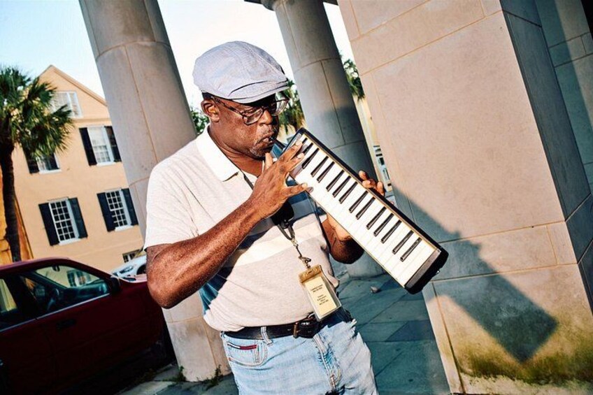 Un hombre tocando un teclado al aire libre, con un coche y edificios al fondo. 5