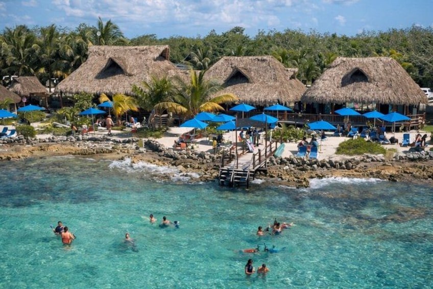 Ein Strand mit klarem türkisblauem Wasser, Hütten mit Reetdach und Menschen beim Schwimmen und Entspannen. 2