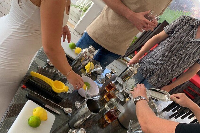 A group of people preparing drinks with various bottles and a yellow measuring cup on a table. 2