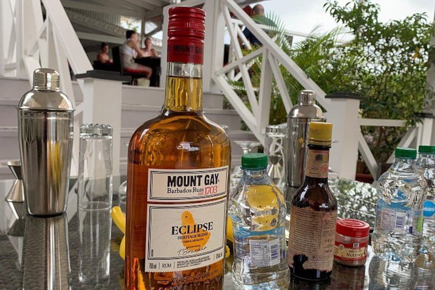 En flaske Mount Gay Barbados rom, en cocktailshaker og diverse bartilbehør på et bord. 2