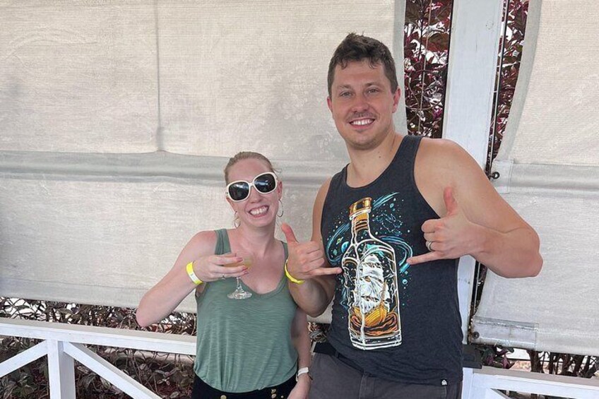 Two people standing on a porch with a white railing, a white tent in the background, and a bottle graphic on the man's tank top. 4