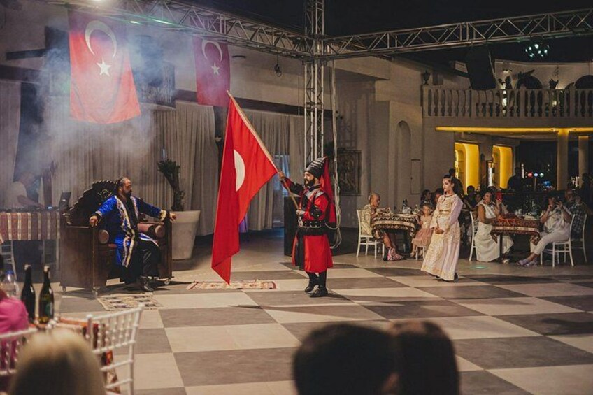 A cultural performance with a flag, a seated figure in traditional attire, and an audience. 2